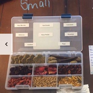 May include: A clear plastic storage box with a variety of dried herbs and flowers. The box is labeled with the names of the herbs and flowers, including Goji Berries, Oat Groats, Calendula, Hibiscus, Rose Petals, Rosehips, Cookies, and Pops.