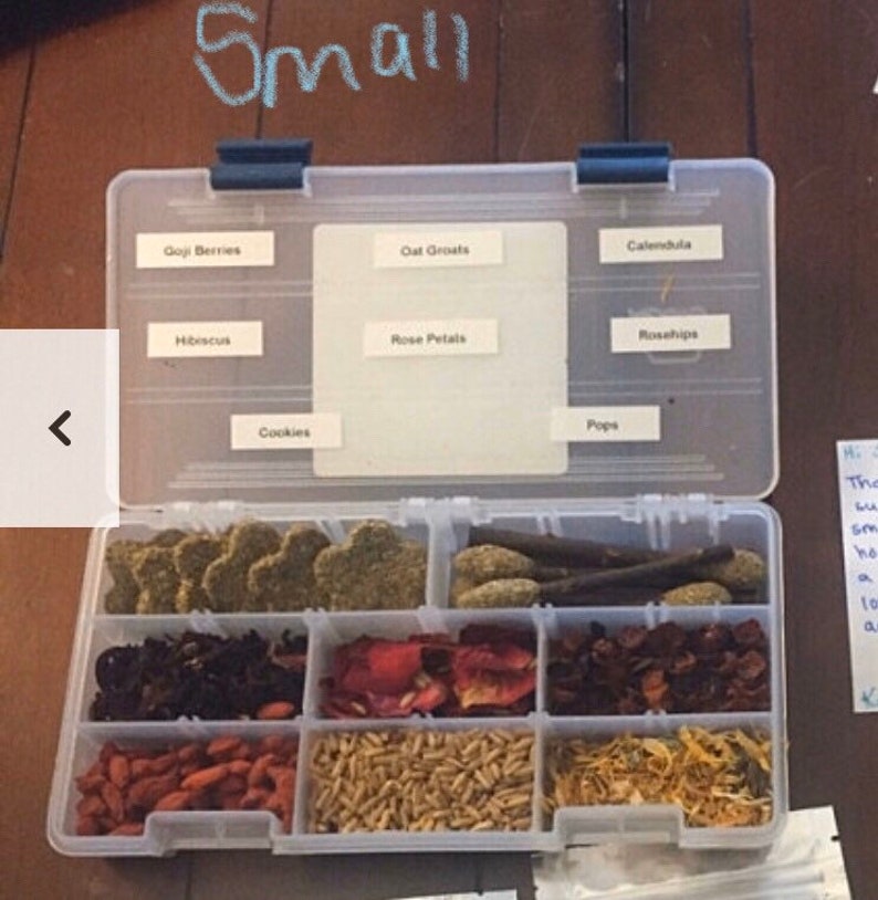 May include: A clear plastic storage box with a variety of dried herbs and flowers. The box is labeled with the names of the herbs and flowers, including Goji Berries, Oat Groats, Calendula, Hibiscus, Rose Petals, Rosehips, Cookies, and Pops.
