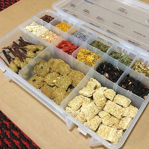 May include: A clear plastic storage container with multiple compartments filled with various dried herbs and other ingredients. The compartments are labeled with the names of the herbs, including "Oat Groats", "Goji Berries", "Lavender", "Peppermint", "Calendula", "Hibiscus", "Dandelion", and "Shredded Wheat".