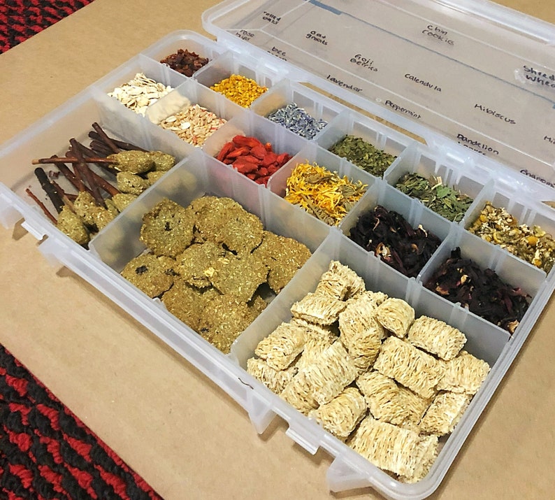 May include: A clear plastic storage container with multiple compartments filled with various dried herbs and other ingredients. The compartments are labeled with the names of the herbs, including "Oat Groats", "Goji Berries", "Lavender", "Peppermint", "Calendula", "Hibiscus", "Dandelion", and "Shredded Wheat".
