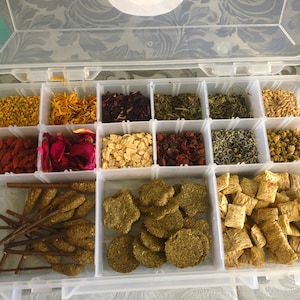 May include: A clear plastic storage container with 16 compartments filled with various dried herbs, flowers, and other ingredients. The container is open and the compartments are visible.