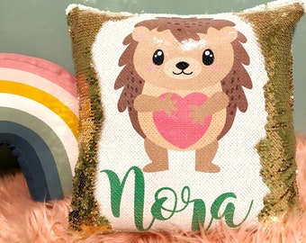 May include: A square sequin pillow featuring a cartoon hedgehog holding a pink heart, with the name Nora printed in green script below. The pillow has gold-colored sequins on the sides and a white center panel. A rainbow-shaped pillow is nearby.
