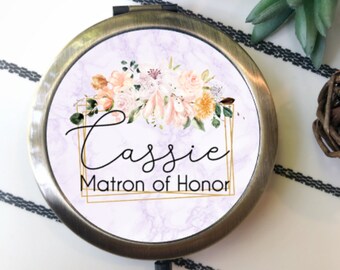 May include: A round, antique brass-toned compact mirror featuring a light purple marble-patterned background. The center displays a floral arrangement in peach and white tones, a gold geometric frame, and the text "Cassie" and "Matron of Honor."