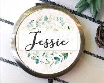 May include: A round, antique brass-toned compact mirror featuring a central white rectangular label with the name Jessie written in black script. The design is surrounded by a watercolor-style wreath of green eucalyptus leaves on a light beige background.