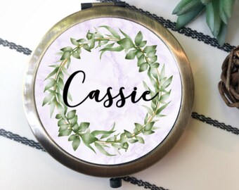 May include: A round, antique brass-toned compact mirror featuring a personalized design on the lid. The center displays the name Cassie in black script, surrounded by a circular wreath of green leaves and a thin gold ring against a light purple marble background.