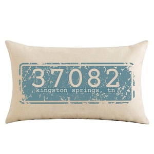 May include: A beige pillow with a blue and white distressed rectangle design. The rectangle has the text "37082" and "kingston springs, tn" printed in white.