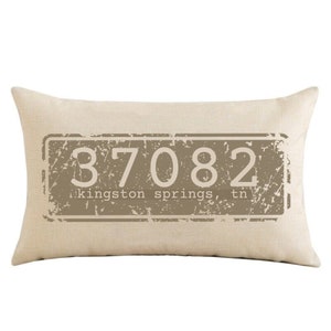 May include: Beige rectangular pillow with a brown distressed rectangle design featuring the text "37082 kingston springs, tn".