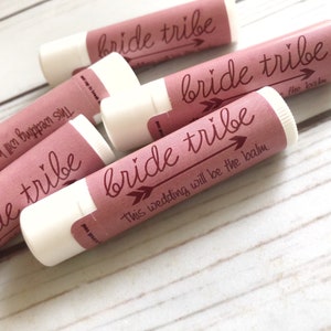 May include: A close-up of several pink lip balm tubes with white caps. The labels on the tubes say "bride tribe" with an arrow pointing to the right. One label says "This wedding will be the balm."