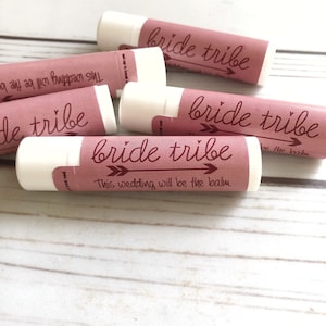 May include: A set of five pink and white lip balm tubes with the text "bride tribe" and an arrow graphic. The text "This wedding will be the balm" is printed on one of the tubes.