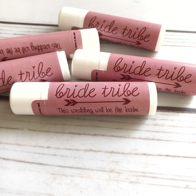 May include: A set of five pink and white lip balm tubes with the text "bride tribe" and an arrow graphic. The text "This wedding will be the balm" is printed on one of the tubes.