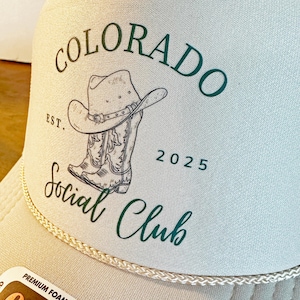 Peut inclure: Casquette de camionneur beige avec un panneau avant blanc et une visi&egrave;re incurv&eacute;e. Le chapeau pr&eacute;sente un graphisme de chapeau de cowboy et de bottes avec les mots "COLORADO Social Club" en vert. L'ann&eacute;e "2025" est &eacute;galement imprim&eacute;e sur le chapeau.