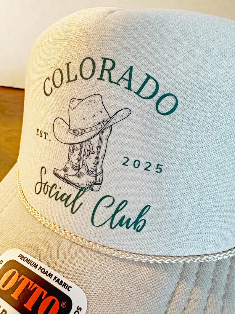 Peut inclure: Casquette de camionneur beige avec un panneau avant blanc et une visi&egrave;re incurv&eacute;e. Le chapeau pr&eacute;sente un graphisme de chapeau de cowboy et de bottes avec les mots "COLORADO Social Club" en vert. L'ann&eacute;e "2025" est &eacute;galement imprim&eacute;e sur le chapeau.