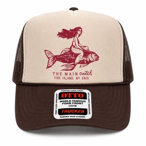 May include: A brown and cream trucker hat featuring a maroon graphic of a person riding a large fish. The text reads THE MAIN catch FIRE ISLAND, NY 2026. A white sticker on the brim displays OTTO WORLD FAMOUS FOAM FRONT OSFM TRUCKER.