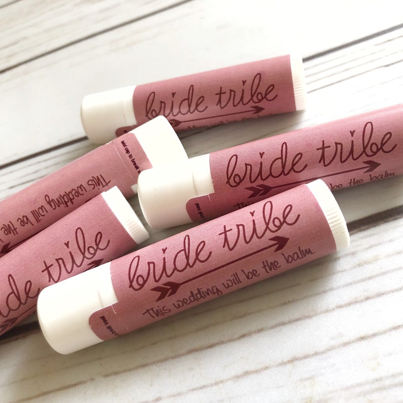 May include: Five pink lip balm tubes with white caps. The labels are decorated with the text "bride tribe" and an arrow. One label reads "This wedding will be the balm."
