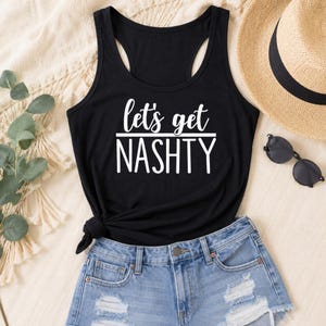 May include: A black racerback tank top featuring the white text "let's get NASHTY" printed on the front. The garment has a scoop neckline and a knotted detail at the bottom hem, paired with light blue distressed denim shorts.