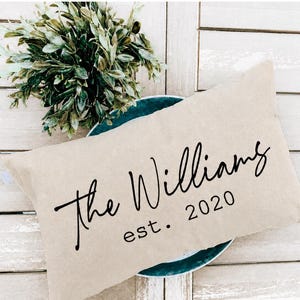 Op de afbeelding: Een rechthoekig beige sierkussen met de zwarte opdruk "The Williams" in een cursief lettertype boven de tekst "est. 2020" in een eenvoudig schreefloos lettertype. Het kussen ligt gedeeltelijk over een donkergroenblauw rond bord heen.