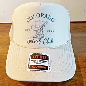 Peut inclure: Casquette de camionneur beige clair avec le texte "COLORADO Social Club" et un dessin d'un chapeau et de bottes de cowboy. Le chapeau a un devant en mousse et une fermeture &agrave; pression. Le chapeau porte &eacute;galement le texte "EST. 2025".