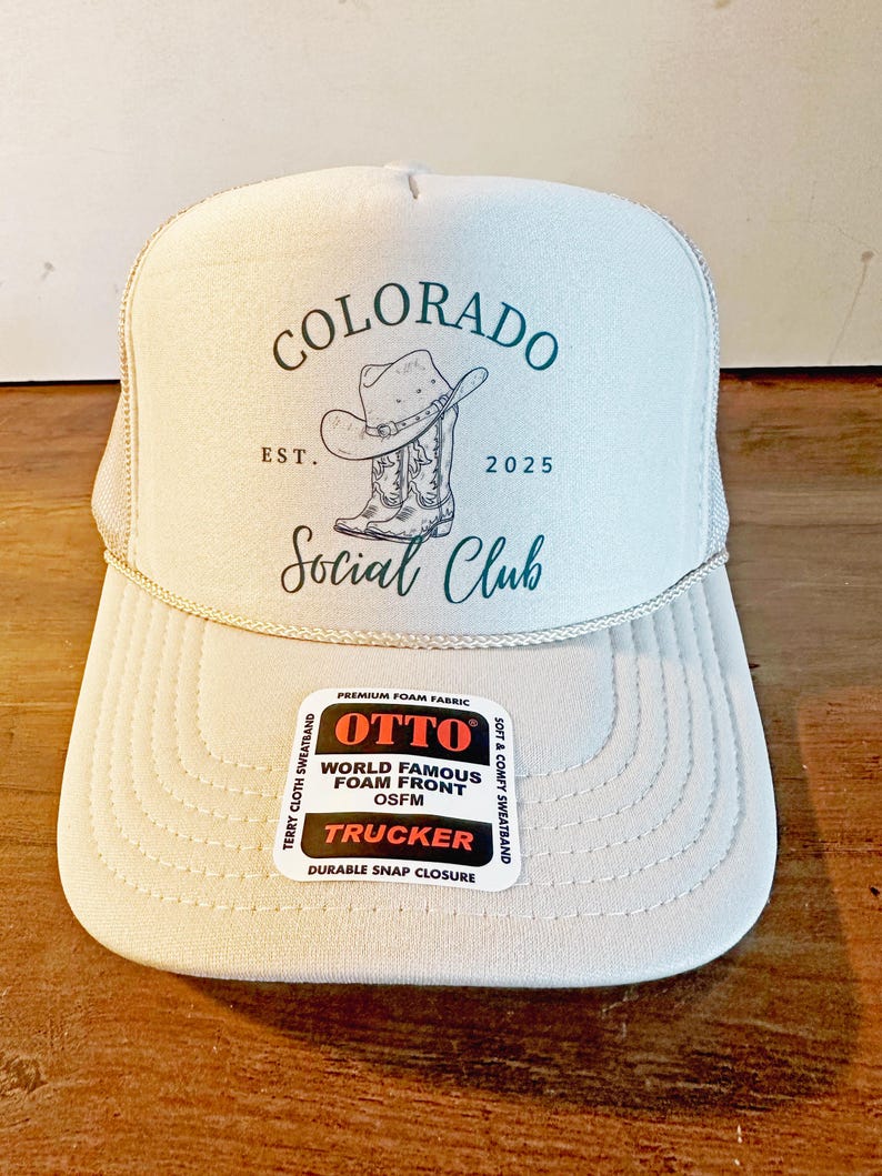 Peut inclure: Casquette de camionneur beige clair avec le texte "COLORADO Social Club" et un dessin d'un chapeau et de bottes de cowboy. Le chapeau a un devant en mousse et une fermeture &agrave; pression. Le chapeau porte &eacute;galement le texte "EST. 2025".