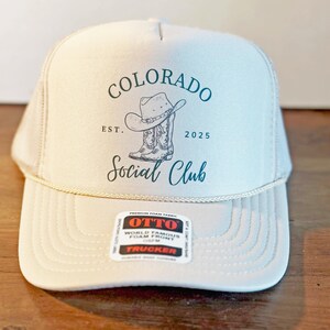 Peut inclure: Casquette de camionneur beige avec un dos en filet et une visi&egrave;re incurv&eacute;e. La casquette porte l'inscription "Colorado Social Club" en script teal, ainsi qu'un graphique de chapeau et de botte de cowboy. La casquette porte &eacute;galement la date "2025".