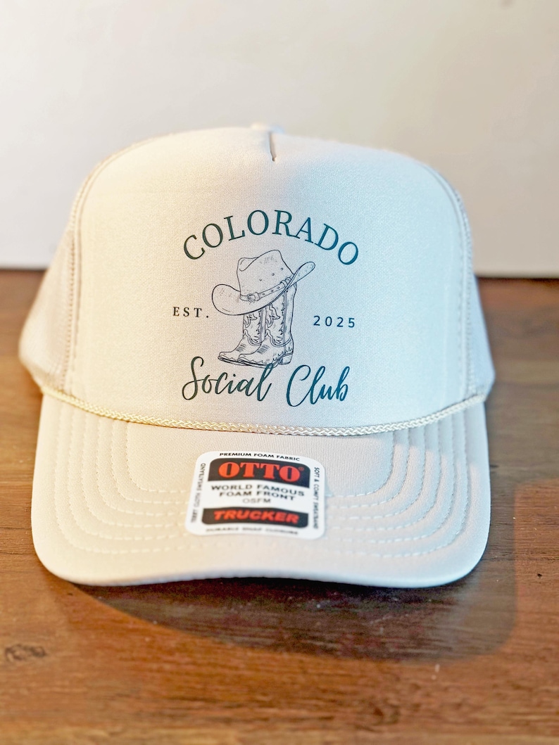 Peut inclure: Casquette de camionneur beige avec un dos en filet et une visi&egrave;re incurv&eacute;e. La casquette porte l'inscription "Colorado Social Club" en script teal, ainsi qu'un graphique de chapeau et de botte de cowboy. La casquette porte &eacute;galement la date "2025".