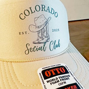 Peut inclure: Casquette de camionneur beige avec l'inscription "COLORADO Social Club" et un dessin de chapeau et de botte de cowboy. La casquette a une visi&egrave;re incurv&eacute;e et une &eacute;tiquette "OTTO TRUCKER".