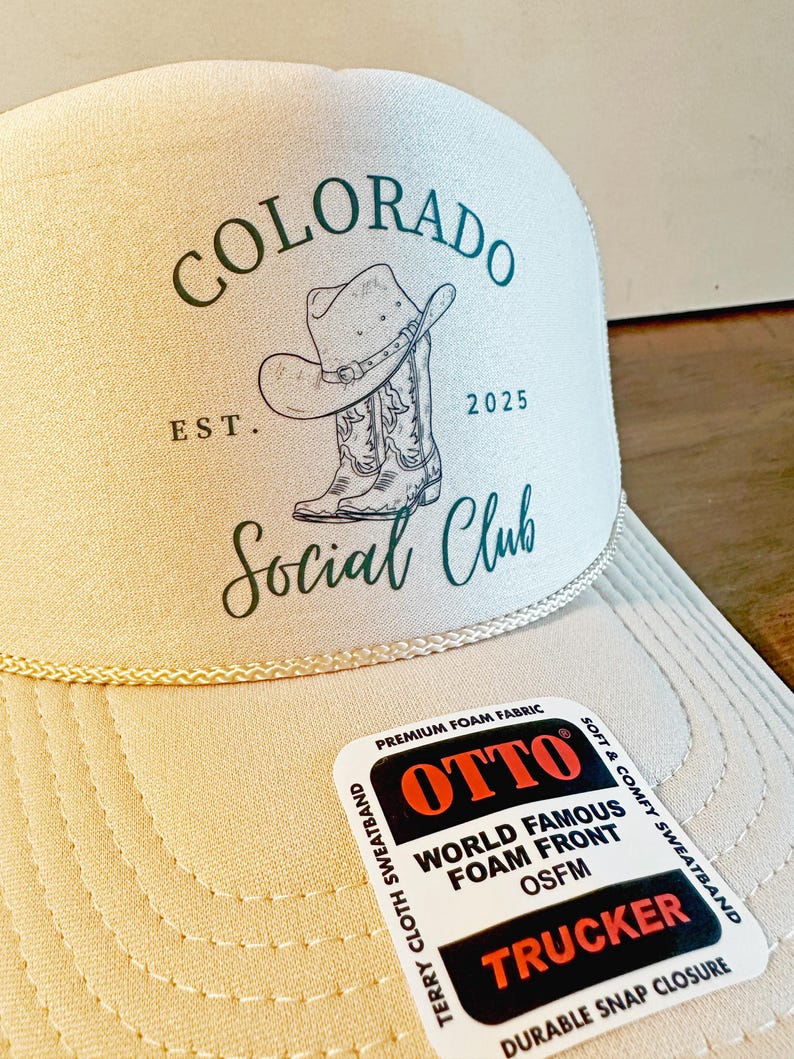 Peut inclure: Casquette de camionneur beige avec l'inscription "COLORADO Social Club" et un dessin de chapeau et de botte de cowboy. La casquette a une visi&egrave;re incurv&eacute;e et une &eacute;tiquette "OTTO TRUCKER".
