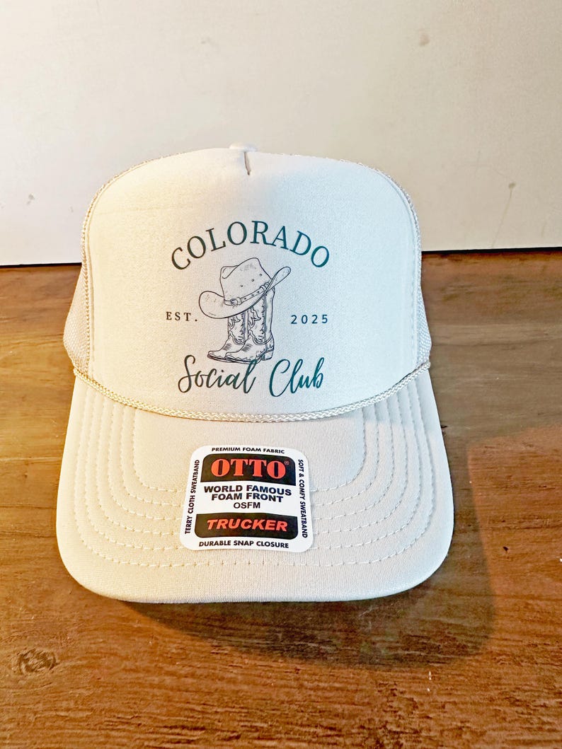 Peut inclure: Casquette de camionneur beige clair avec un dos en filet blanc et une visi&egrave;re incurv&eacute;e. Le devant du chapeau porte l'inscription "COLORADO Social Club" avec un dessin de chapeau et de botte de cowboy. Le chapeau a une &eacute;tiquette "OTTO".