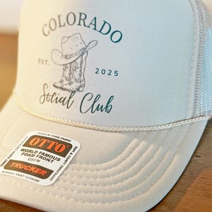 Peut inclure: Casquette de camionneur gris clair avec un dos en filet blanc. Le chapeau porte les mots "COLORADO Social Club" en script vert d'eau, ainsi qu'un dessin de chapeau et de botte de cowboy. Le chapeau a un d&eacute;tail de corde blanche et une &eacute;tiquette "OTTO".