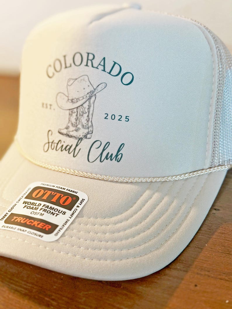 Peut inclure: Casquette de camionneur gris clair avec un dos en filet blanc. Le chapeau porte les mots "COLORADO Social Club" en script vert d'eau, ainsi qu'un dessin de chapeau et de botte de cowboy. Le chapeau a un d&eacute;tail de corde blanche et une &eacute;tiquette "OTTO".