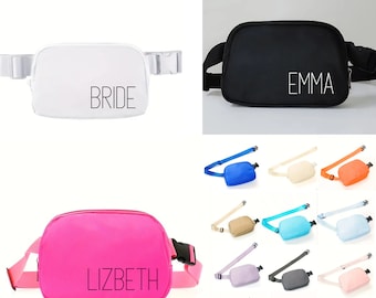 May include: A collection of personalized fanny packs in various colors, including white, black, and pink. Each pack features a name or word printed on the front, such as "BRIDE," "EMMA," and "LIZBETH." The packs have adjustable straps and a zippered main compartment.