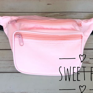 May include: A pink fanny pack with two zippered compartments and a black strap. The fanny pack has the text "SWEET PEA" with hearts on either side.