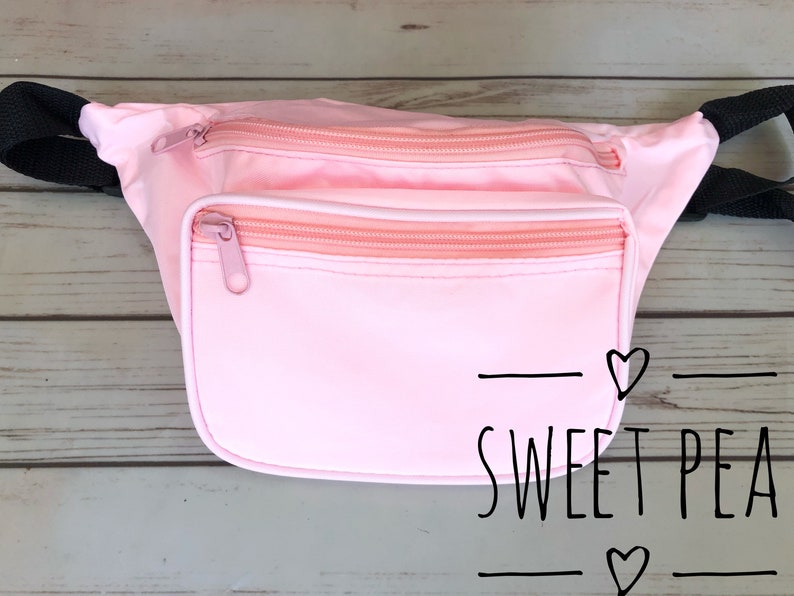May include: A pink fanny pack with two zippered compartments and a black strap. The fanny pack has the text "SWEET PEA" with hearts on either side.