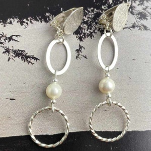 May include: A pair of silver clip-on earrings featuring a pearl and three silver rings. The rings are of varying sizes and shapes.