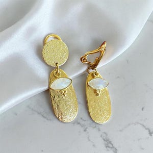 Peut inclure: Une paire de boucles d'oreilles dorées. Chaque boucle d'oreille présente un pendentif ovale allongé texturé avec une pierre blanche de forme ovale. Les boucles d'oreilles ont une pièce supérieure circulaire et une fermeture de type clip.
