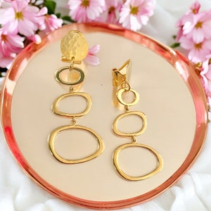 Peut inclure: Une paire de boucles d'oreilles pendantes dorées avec un disque martelé et trois maillons ovales. Les boucles d'oreilles sont sur un plateau rose et or.
