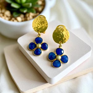 Peut inclure: Boucles d'oreilles dorées avec un disque martelé et un motif de fleur bleue. La fleur a quatre pétales et un centre doré. Les boucles d'oreilles sont présentées sur une surface blanche.
