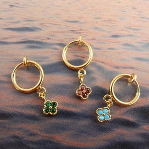 Può includere: Tre orecchini a cerchio dorati con un ciondolo pendente. Ogni ciondolo è un quadrifoglio con una pietra preziosa di colore diverso al centro: rosso, verde e blu.