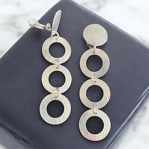 May include: Silver clip-on earrings with a textured finish. The earrings feature four connected circles, each with a smaller circle cut out in the center.