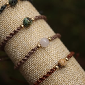 May include: Four bracelets with different coloured stones and gold beads on a beige fabric background. The bracelets are made of brown, dark brown, and green cord.