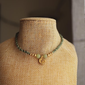 Peut inclure: Un collier tressé vert avec un pendentif soleil doré et de petites perles dorées. Le collier comporte deux perles vert clair et est présenté sur un mannequin beige. Le collier est un bijou.