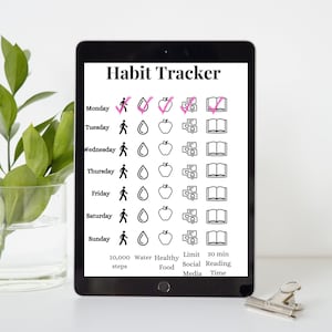 May include: A black tablet displaying a "Habit Tracker" with daily goals. The tracker includes icons for walking, water intake, healthy food, social media limits, and reading time. A silver clip rests on the table.
