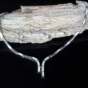 May include: A silver-toned necklace with a geometric design. The necklace features rectangular and square shapes with white inlays. The pendant hangs down in a Y-shape. The necklace is displayed on a piece of wood against a black background.