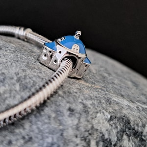 Könnte beinhalten: Ein silbernes Armband mit einem blauen und silbernen Anhänger in Form eines kleinen Gebäudes mit einer Kuppel. Der Anhänger befindet sich an einem silbernen Schlangenkettenarmband.