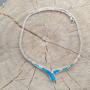 May include: A silver-toned necklace with a geometric design and blue gemstone accents. The necklace has a delicate chain with a repeating pattern and a Y-shaped pendant with vibrant blue stones. The jewelry is displayed on a rustic wooden surface.