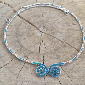 May include: A silver necklace with a blue opal pendant featuring a double spiral design. The necklace has a chain with a geometric pattern.
