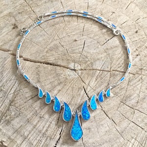 May include: A silver necklace with a teardrop-shaped blue opal pendant and smaller blue opal teardrop accents. The necklace is on a wooden surface.
