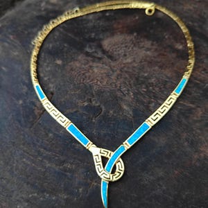 May include: A gold-toned necklace with a geometric design and turquoise blue accents. The necklace features a Greek key pattern and a pendant with a teardrop shape. The jewelry is displayed on a dark wooden surface.