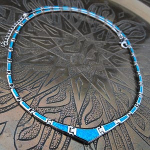 May include: A silver-toned necklace with rectangular and triangular blue opal inlays. The necklace has a geometric design and a delicate chain. The necklace is displayed on a textured, bronze-colored surface.