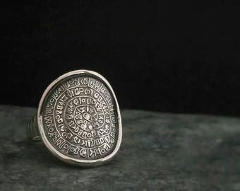 May include: A silver ring with an oval face featuring an engraved circular design with an ancient script. The ring is set on a dark, textured surface, with a black background. The ring's design is detailed and intricate.
