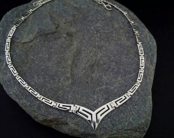 May include: A silver necklace with a geometric pattern. The necklace has a V-shaped pendant with a repeating pattern.